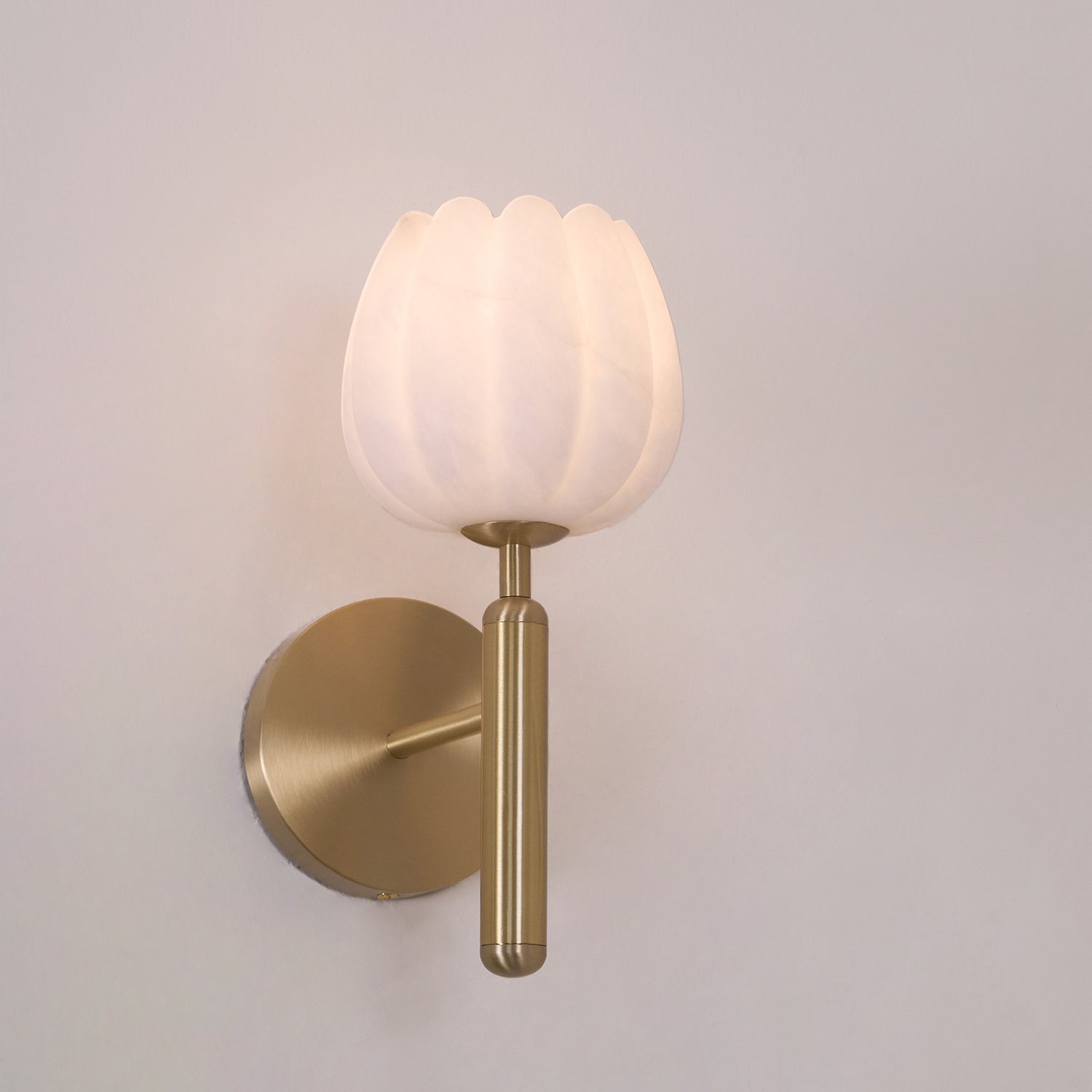 Brass sconce lit on a pale greige wall, front view with a soft glow spilling over the top of the stone petals.