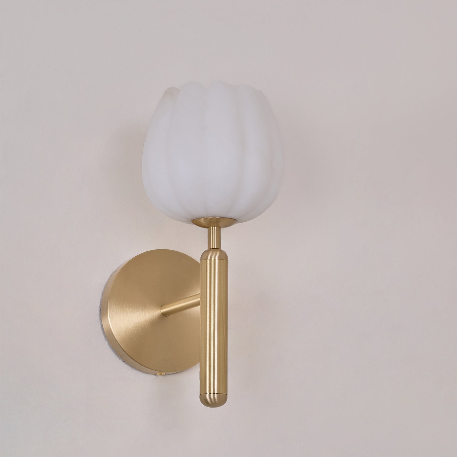 Eurzorion wall light seen front-on against a cream wall, fluted white shade above a ribbed brass stem