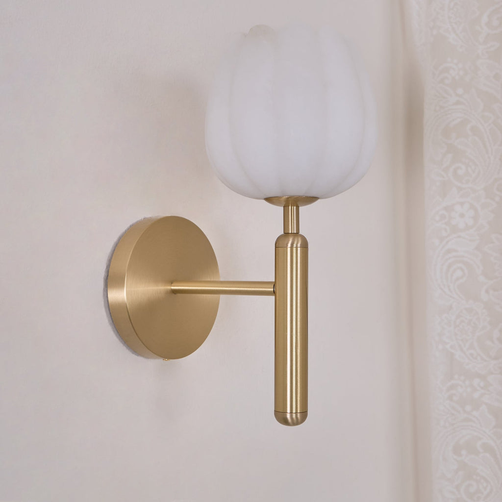 Unlit brass sconce viewed from the side against a cream wall and lace curtain, the shade a flat matte white.