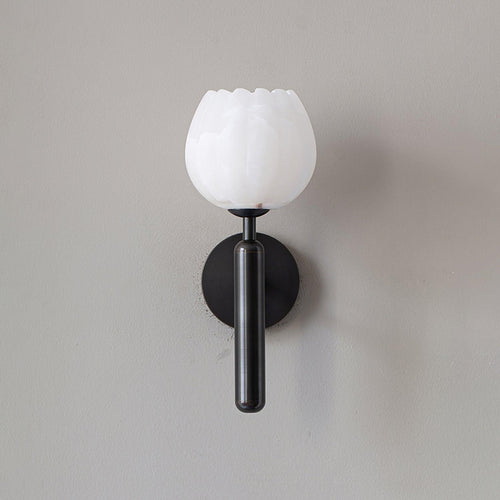 The black-finish sconce unlit on a grey wall, its white alabaster bloom set against the dark metal stem.
