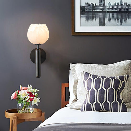 Black-finish sconce lit beside a bed on a dark grey wall, above a bedside table with fresh flowers.