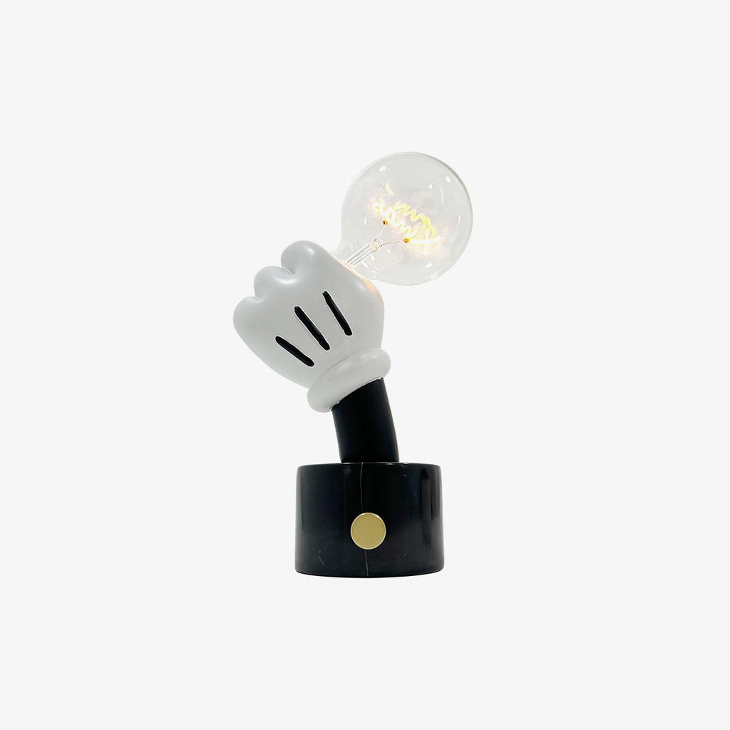 A white glove-shaped hand holds a clear filament bulb above this Clérachic novelty table lamp's black base, set on plain white.