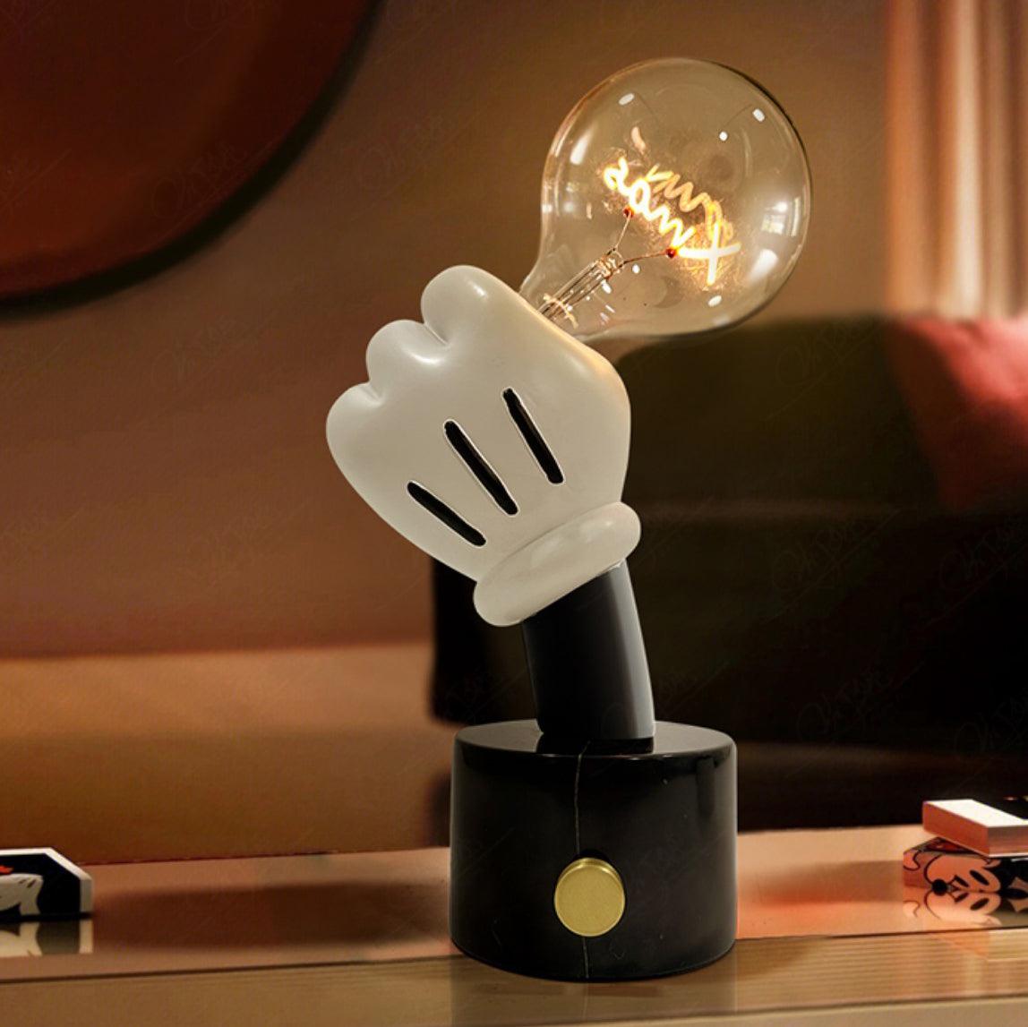 Clérachic novelty table lamp glowing on a low table in a dim room, its white glove hand lifting a clear filament bulb.