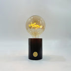 Close-up of the Clérachic novelty lamp's cognac-tinted bulb on its black marble base, the script filament glowing warmly.