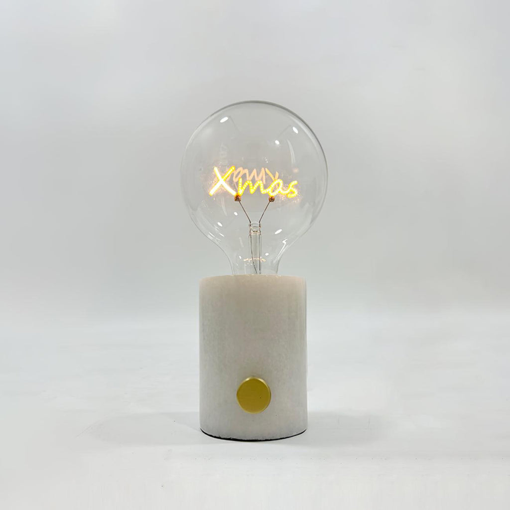 Close-up of the Clérachic novelty lamp's clear bulb on a white marble base, its filament shaped like the word Xmas.