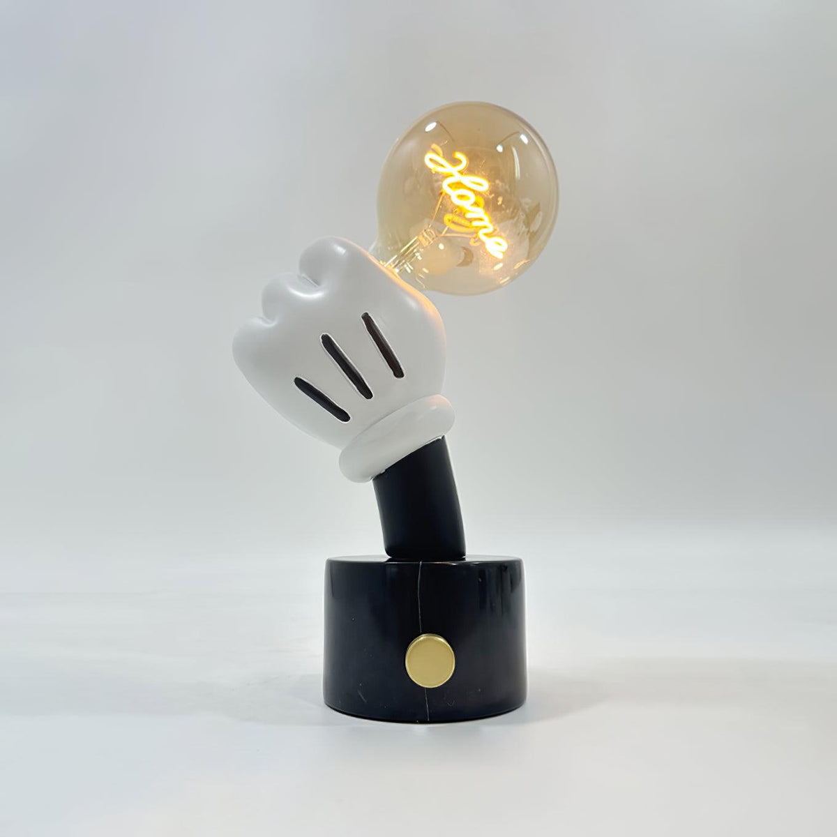 A script-shaped filament glows inside the bulb held by this Clérachic novelty table lamp's glove hand, on black marble against plain grey.