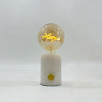 The word Home glows in the filament of this Clérachic novelty lamp's cognac-tinted bulb, set on a white marble base.