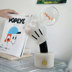 Clérachic novelty table lamp lit on a glass table beside a stack of magazines, a hand lifting the top copy.