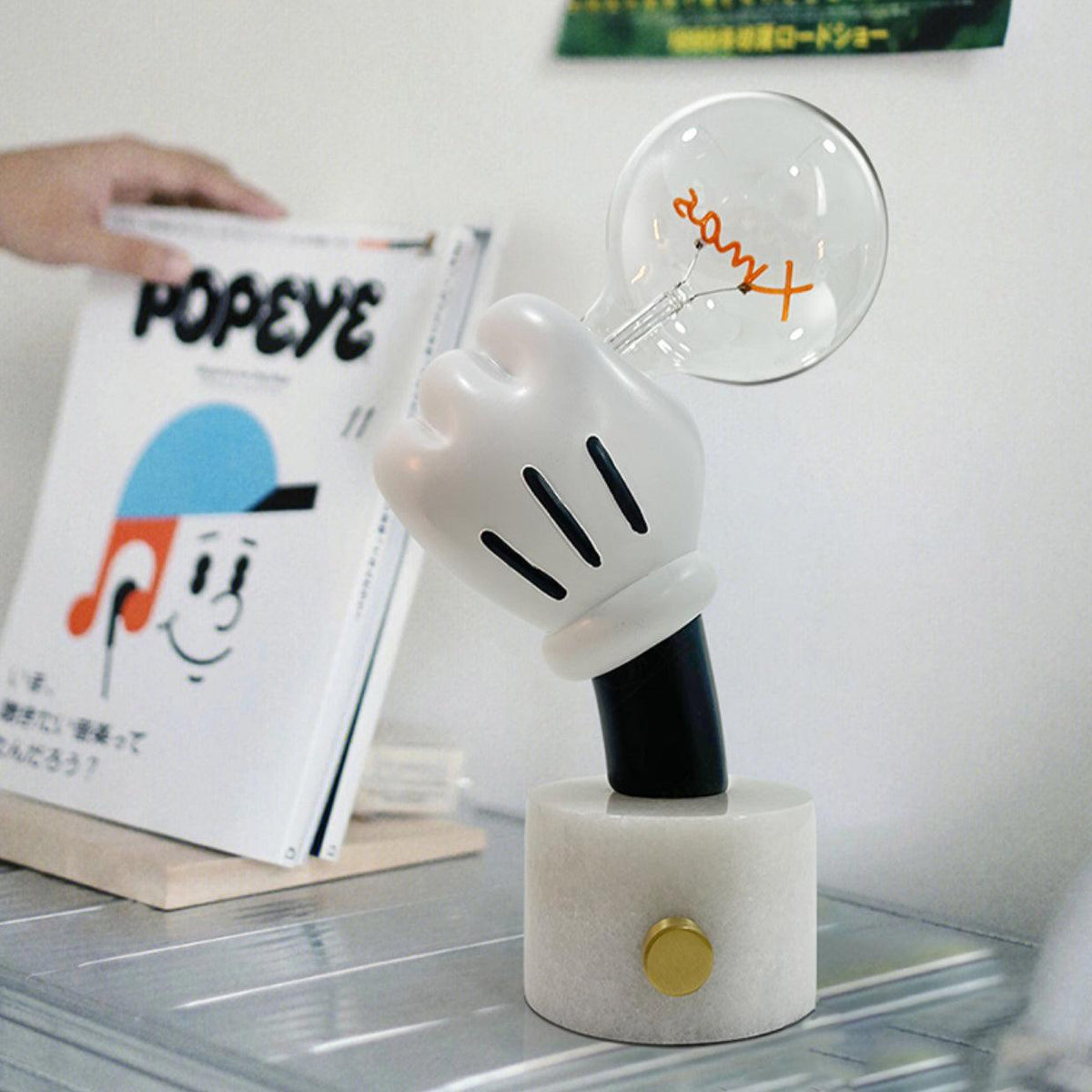Clérachic novelty table lamp lit on a glass table beside a stack of magazines, a hand lifting the top copy.