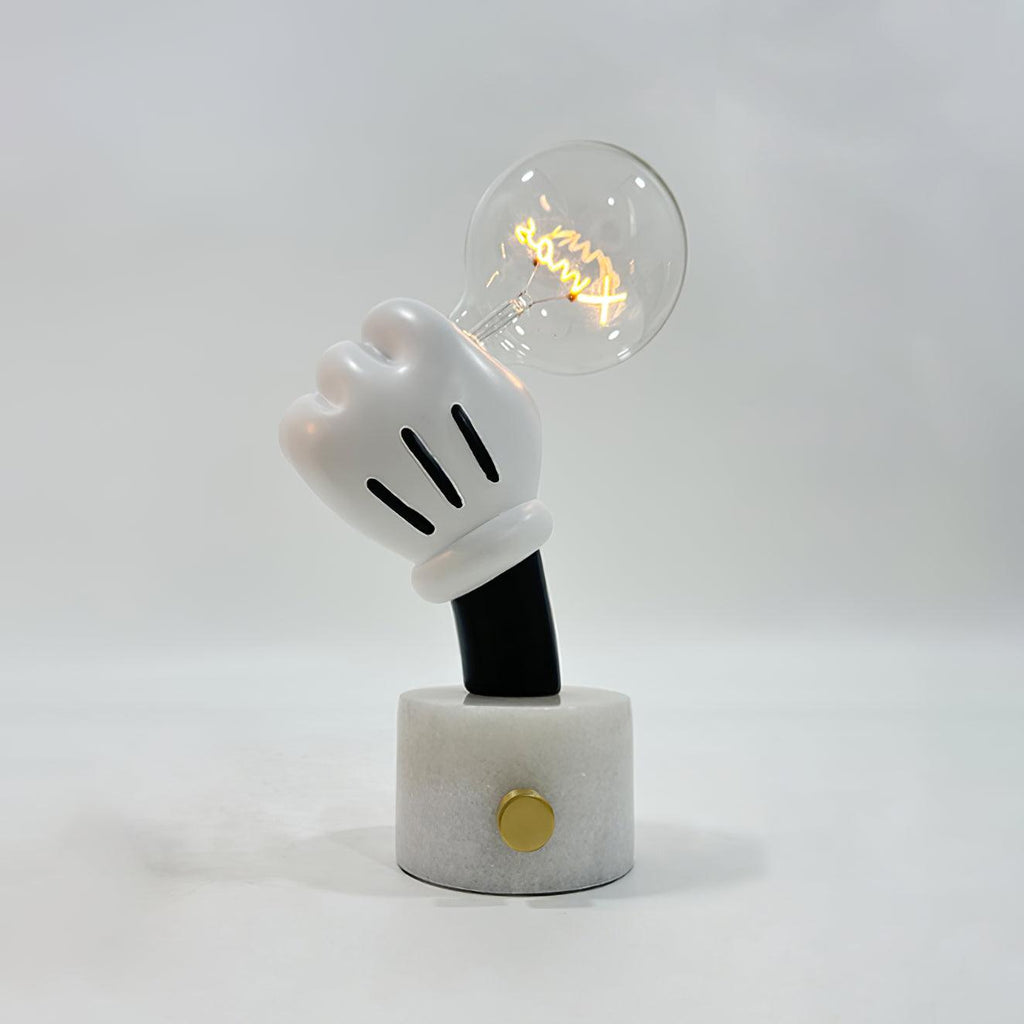 Lifting a lit bulb, the glove-shaped hand tops this Clérachic novelty table lamp on its white marble base and plain grey backdrop.