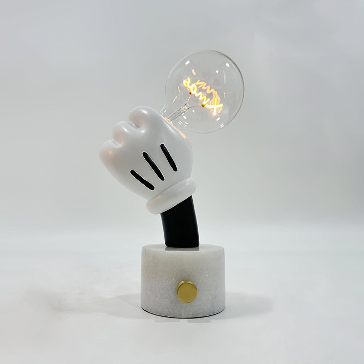 Lifting a lit bulb, the glove-shaped hand tops this Clérachic novelty table lamp on its white marble base and plain grey backdrop.
