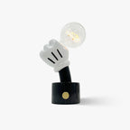 Clérachic novelty table lamp on a plain grey background, a white glove-shaped hand holding a lit Edison bulb above a black marble base.