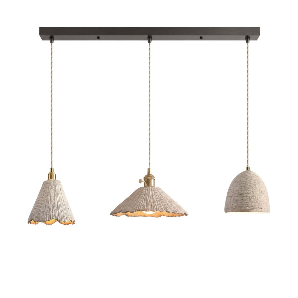 Three microcement pendants on a dark linear ceiling canopy: a tall scalloped cone, a wide scalloped cone and a small bell dome, each on a braided cord