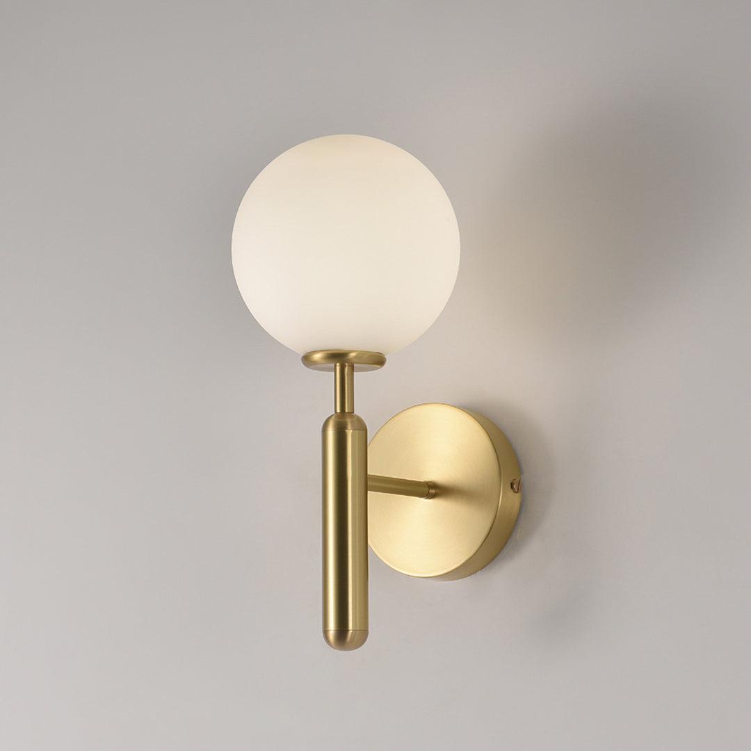 On a plain grey wall, this brass Clérachic wall light shows its round white glass globe and slender arm.