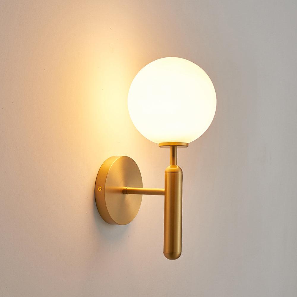A closer view of this brass Clérachic wall light shows its lit white glass globe against a plain off-white wall.