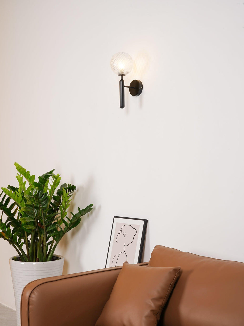 Seen from across the room, a black Clérachic wall light glows softly beside a leafy plant and a framed line drawing.