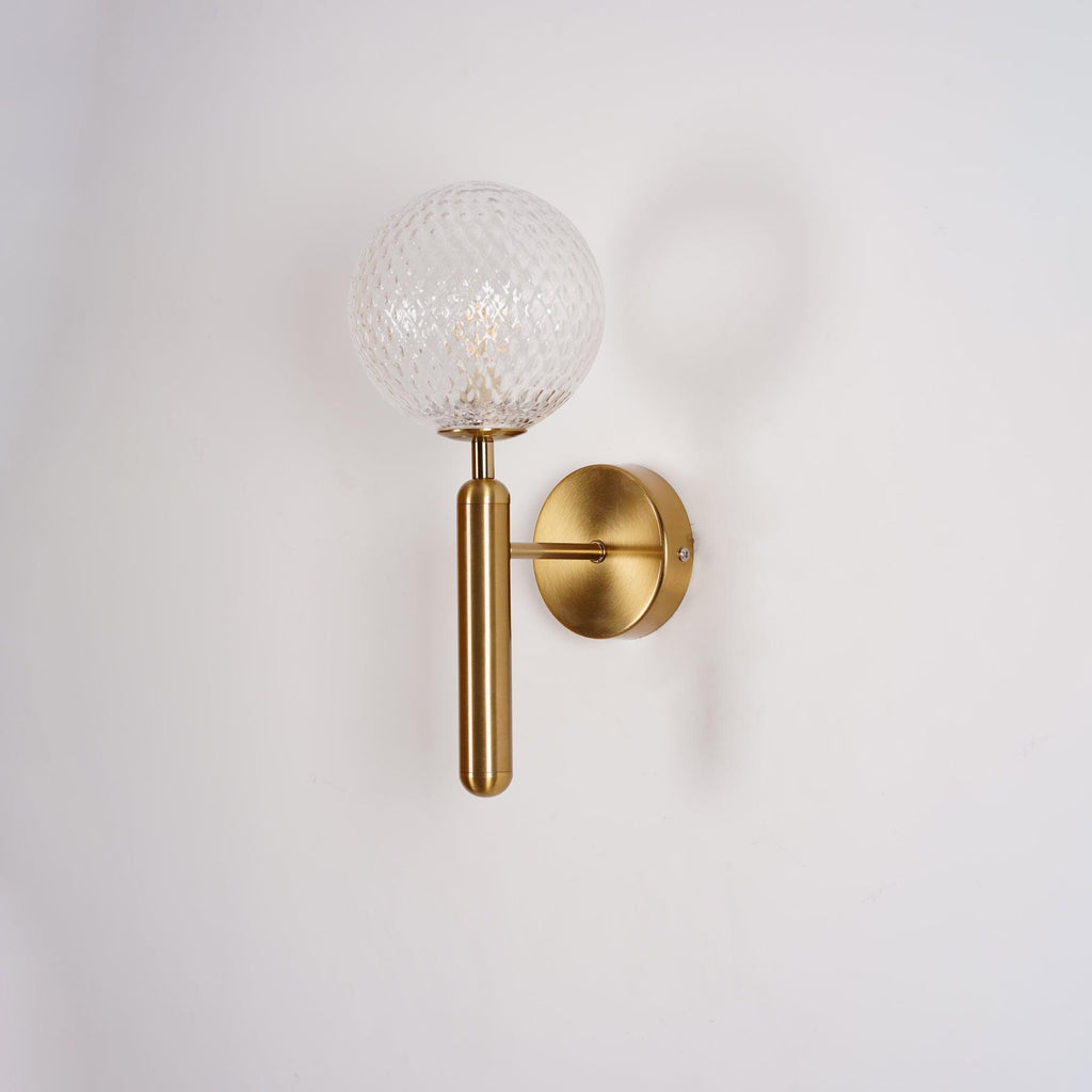 Softly shadowed, this brass Clérachic wall light's dimpled clear glass globe sits against a plain white wall.