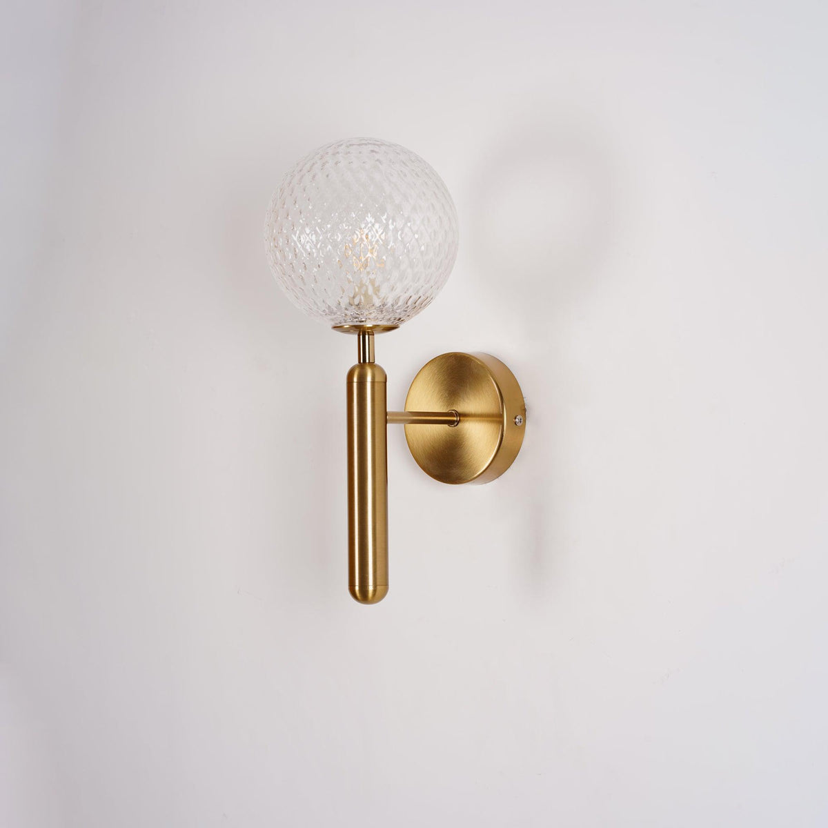 Softly shadowed, this brass Clérachic wall light's dimpled clear glass globe sits against a plain white wall.