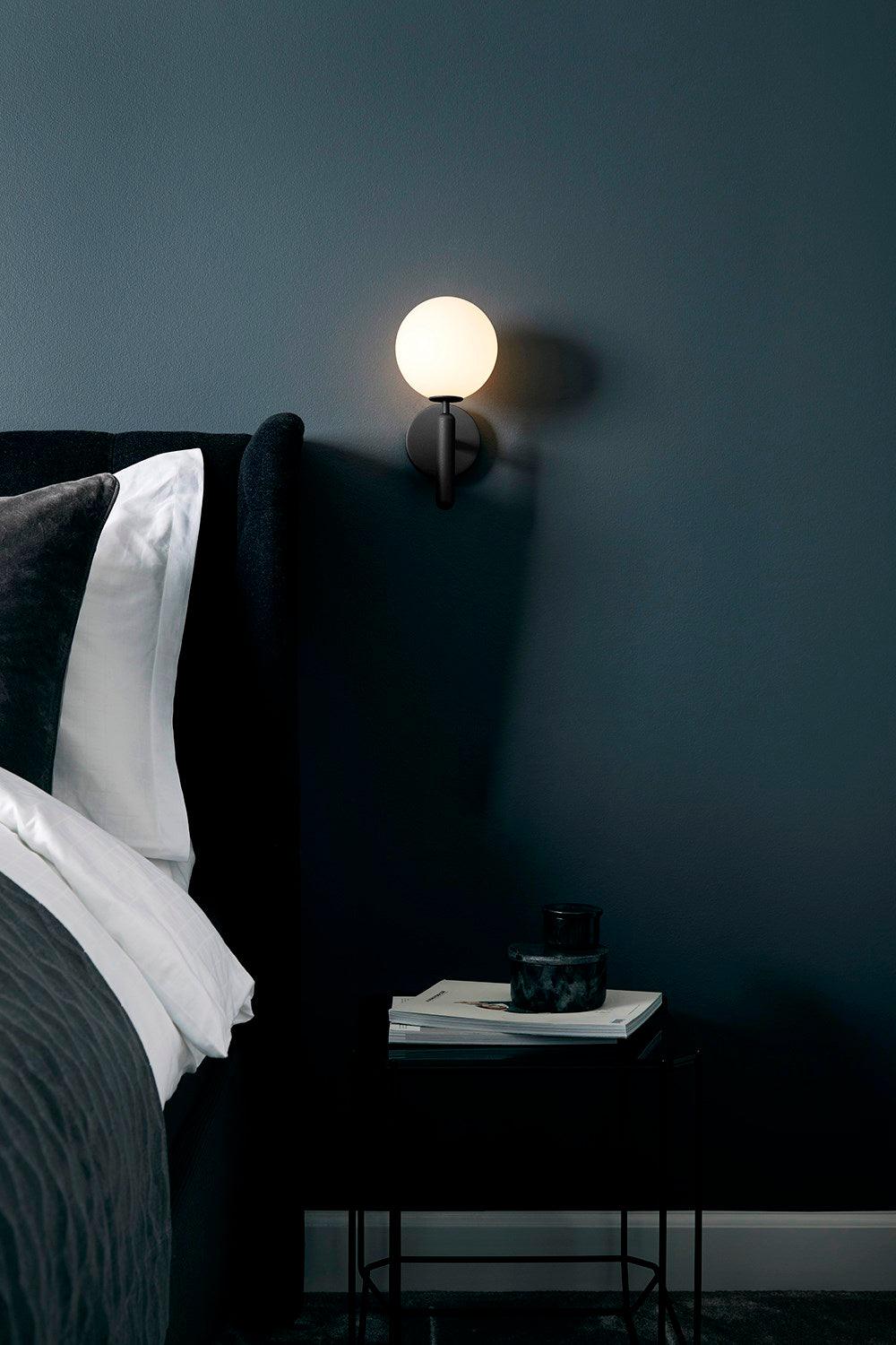 In a deep teal bedroom, this black Clérachic wall light glows above a nightstand with books and a candle.