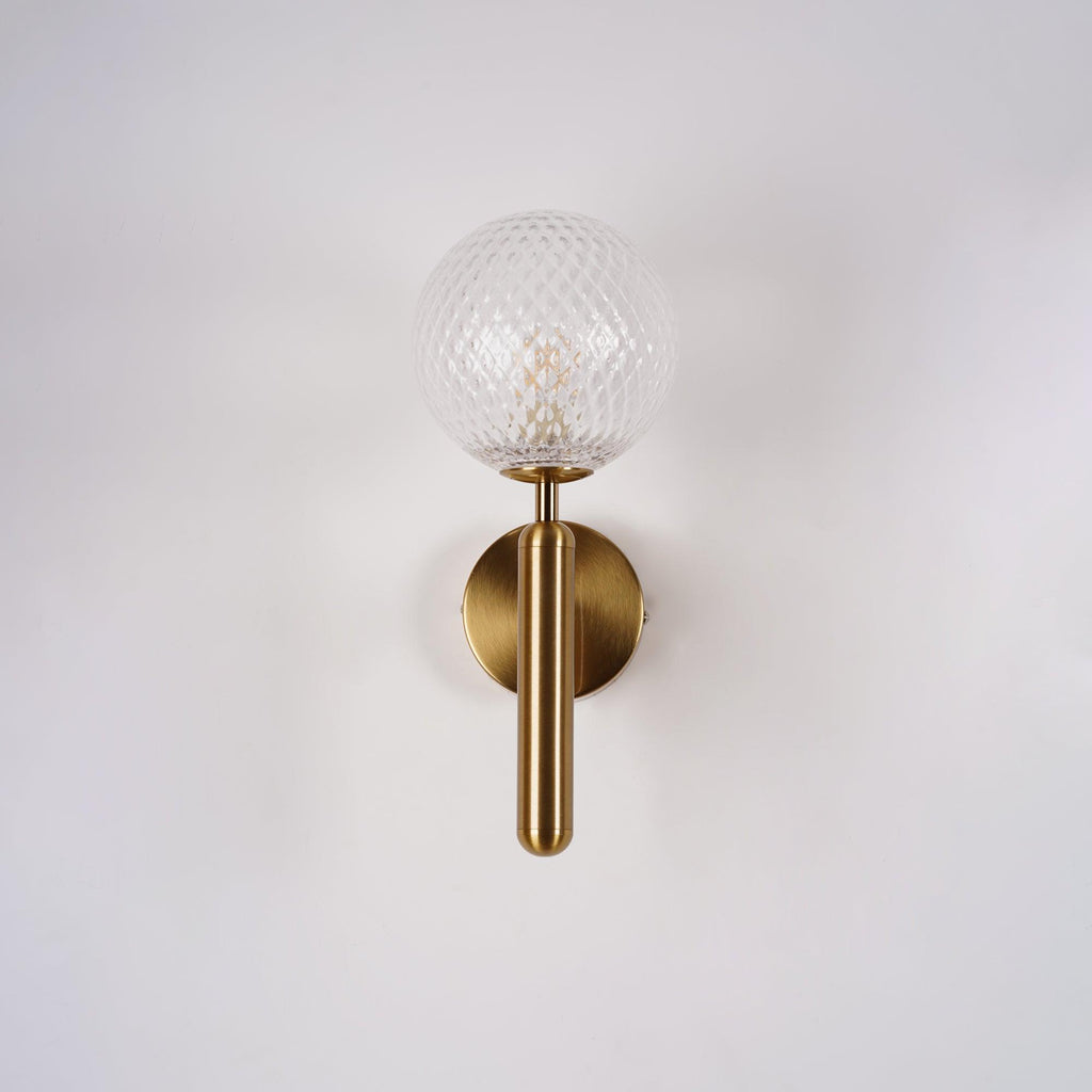 This brass Clérachic wall light shows its dimpled clear glass globe and slender arm against a plain white wall.
