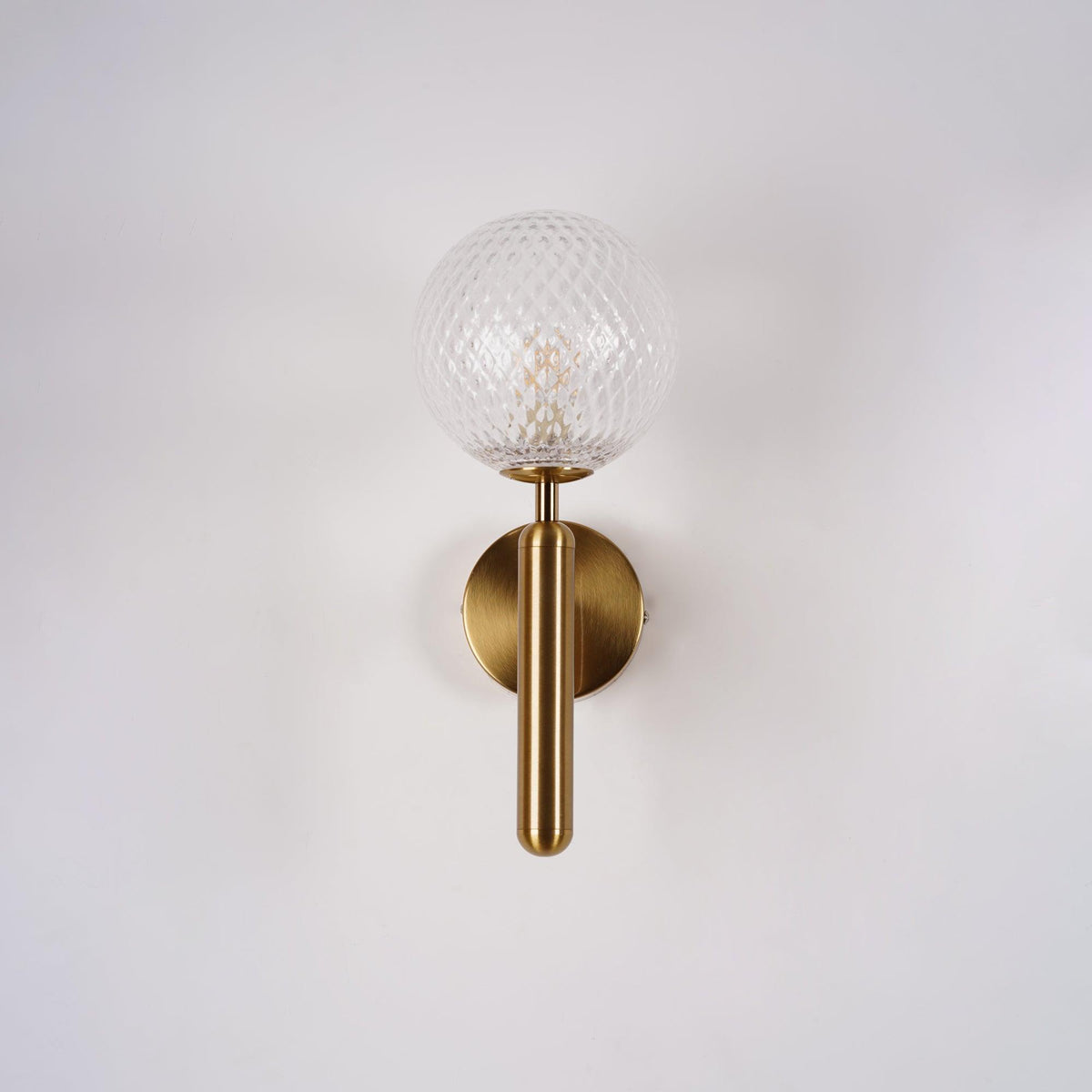 This brass Clérachic wall light shows its dimpled clear glass globe and slender arm against a plain white wall.