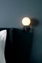 Beside a dark corner of a bedroom, this black Clérachic wall light glows on a deep teal wall near the bed.