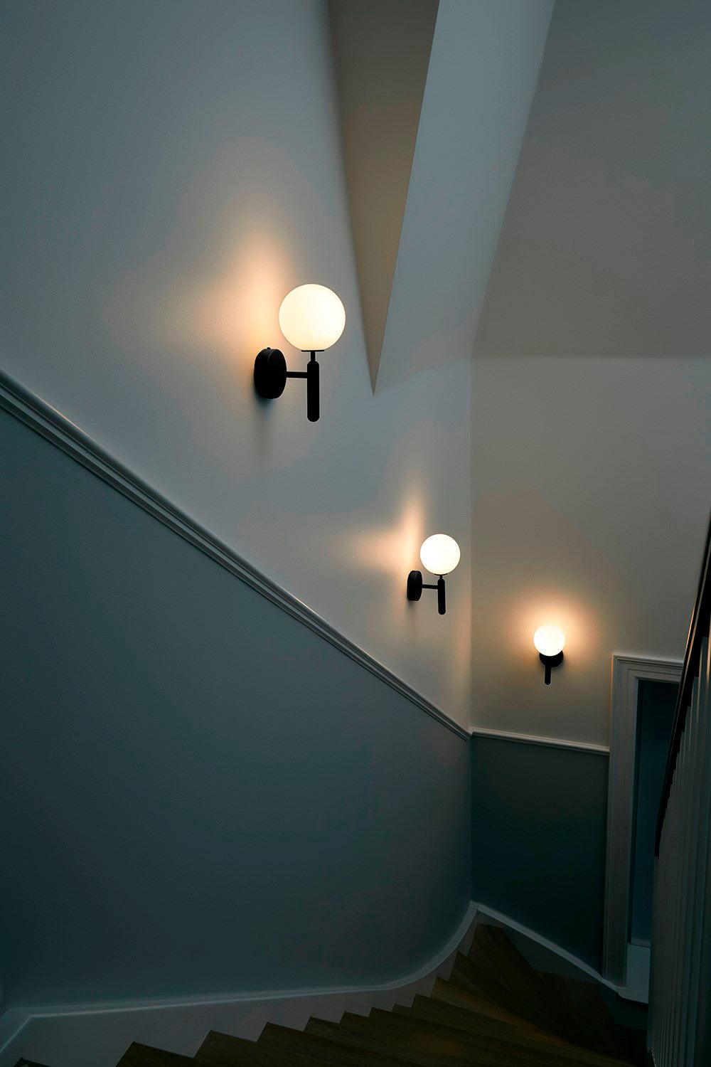 On a stairwell landing, two black Clérachic wall lights with glowing white globes light the way down the stairs.
