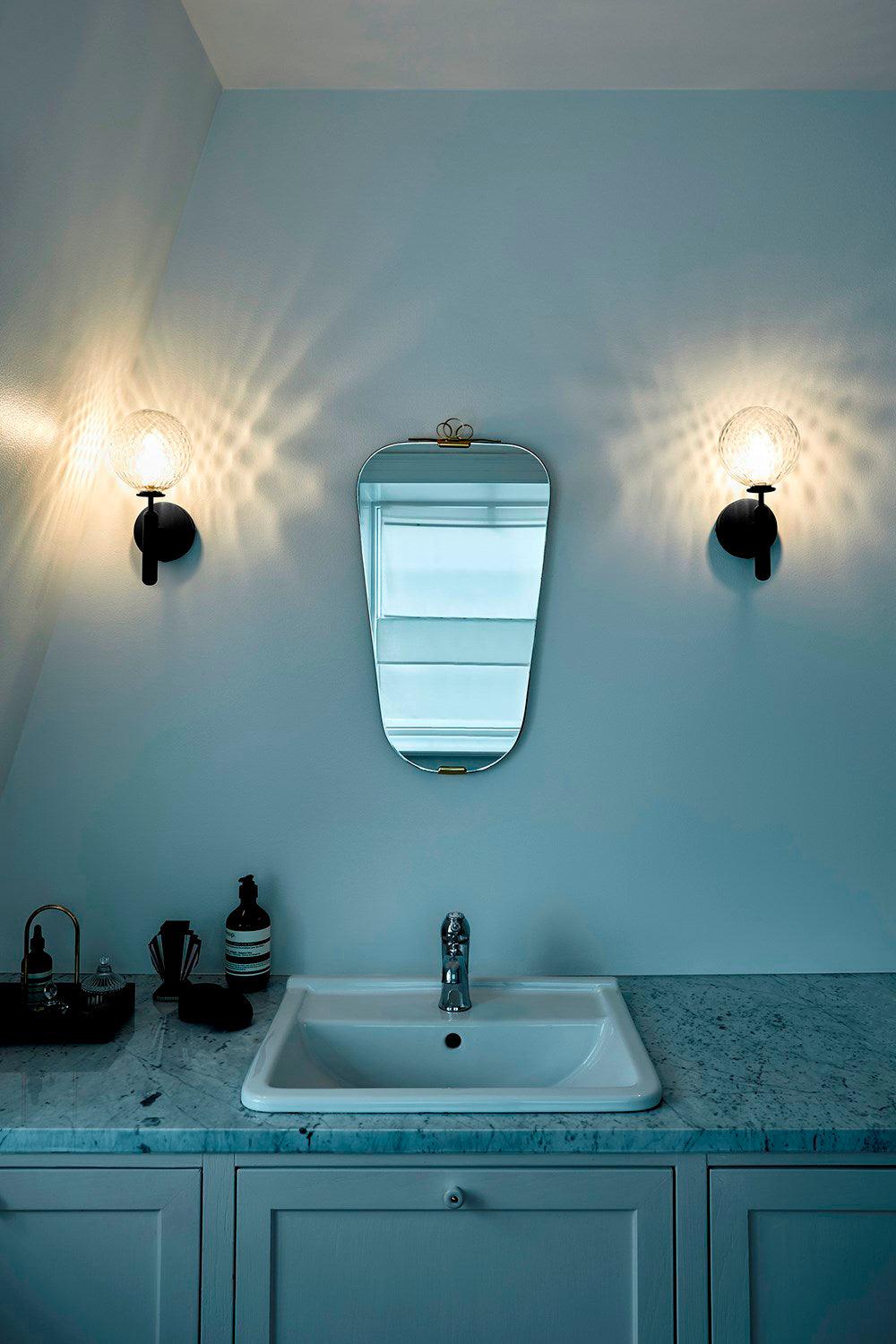 Either side of a mirror, two black Clérachic wall lights with dimpled clear glass globes light a teal bathroom with a marble vanity.