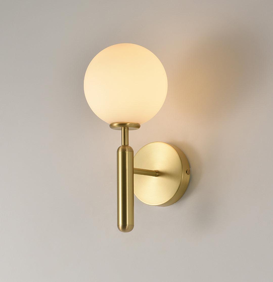 This brass Clérachic wall light with a lit white glass globe stands against a plain cream-coloured wall.