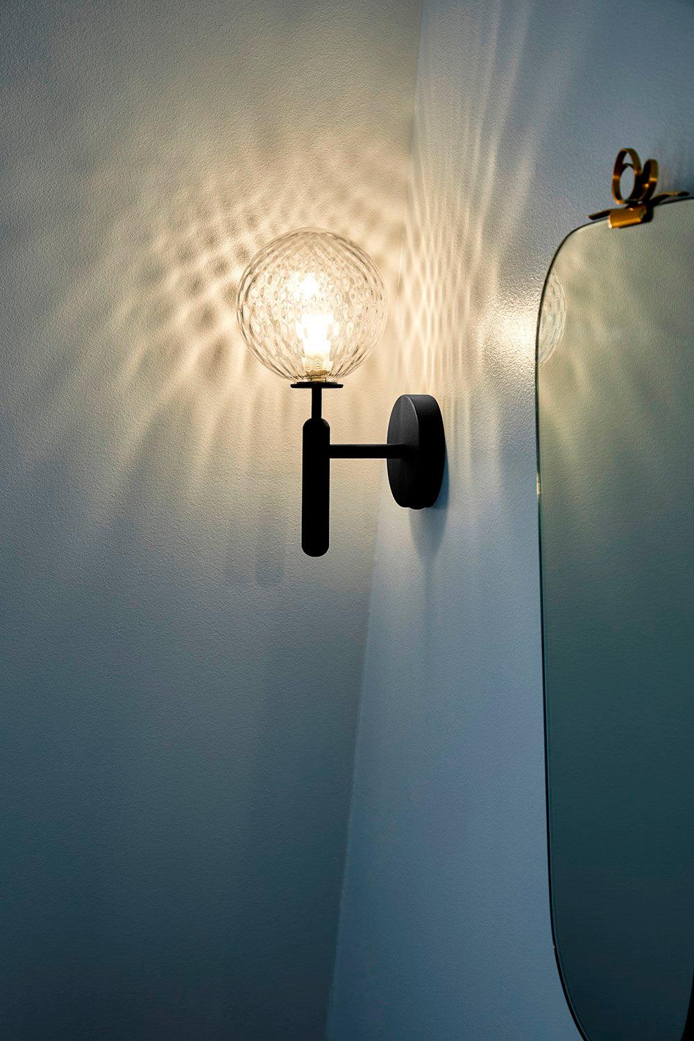 Near the edge of a mirror, this black Clérachic wall light's dimpled clear glass globe casts light across a blue-teal wall.