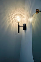 Near the edge of a mirror, this black Clérachic wall light's dimpled clear glass globe casts light across a blue-teal wall.