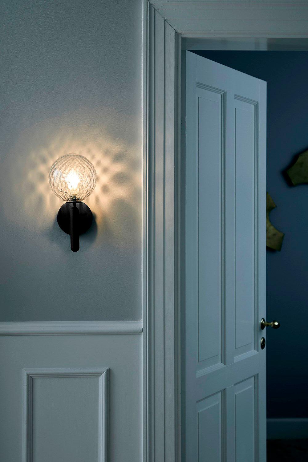 Lit strongly, this black Clérachic wall light's dimpled clear glass globe casts a bright pattern on a blue-grey panelled wall.