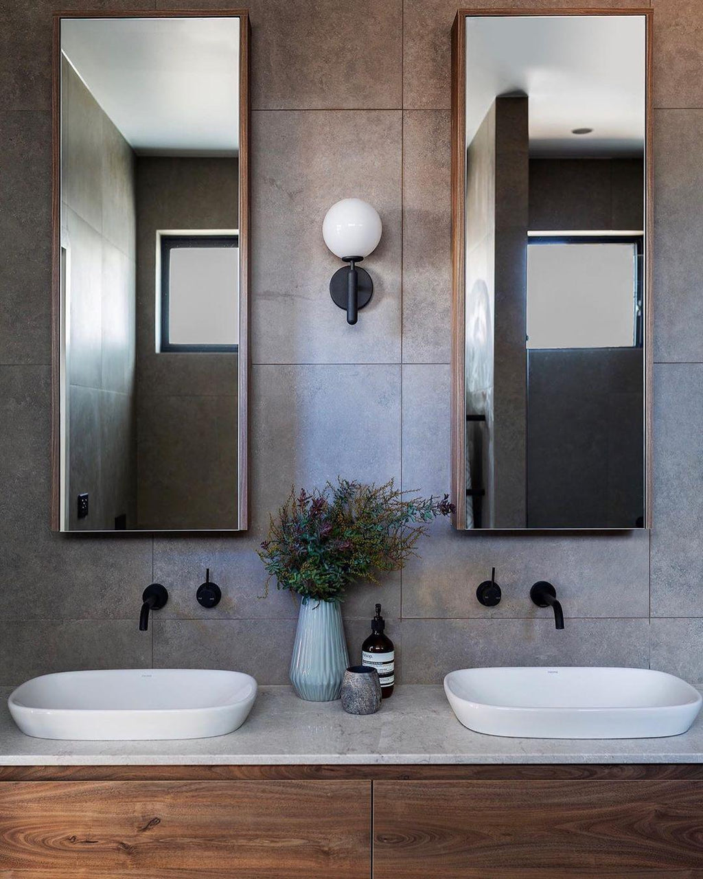 Between two mirrors above vessel sinks with a black tap, this black Clérachic wall light lights a wood-fronted bathroom vanity.