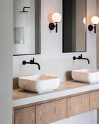 Two black Clérachic wall lights glow between mirrors above two rectangular stone sinks and a wood-fronted vanity.