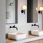 Between two mirrors above a marble vessel sink with a black tap, this black Clérachic wall light glows in a bathroom.
