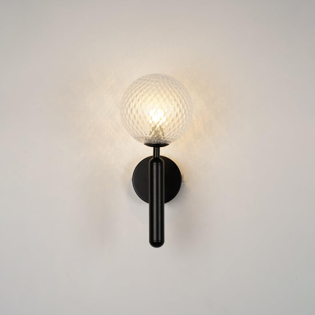 This black Clérachic wall light's dimpled clear glass globe glows gently against a plain grey wall.