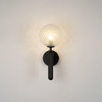 This black Clérachic wall light's dimpled clear glass globe glows gently against a plain grey wall.