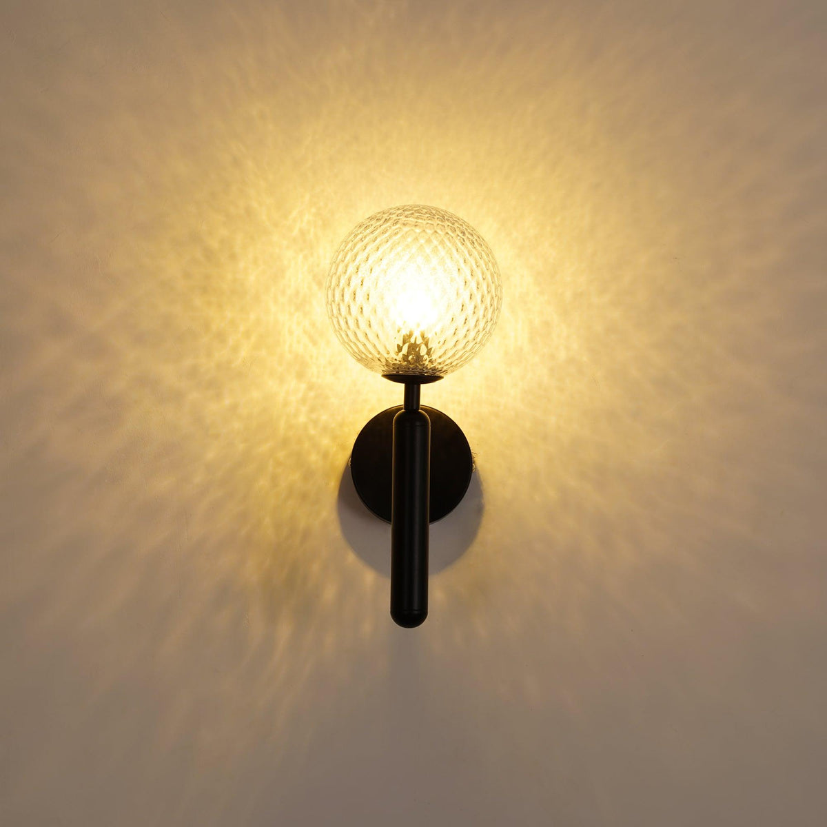 Straight-on, this black Clérachic wall light's dimpled clear glass globe glows warmly, casting light across the wall.