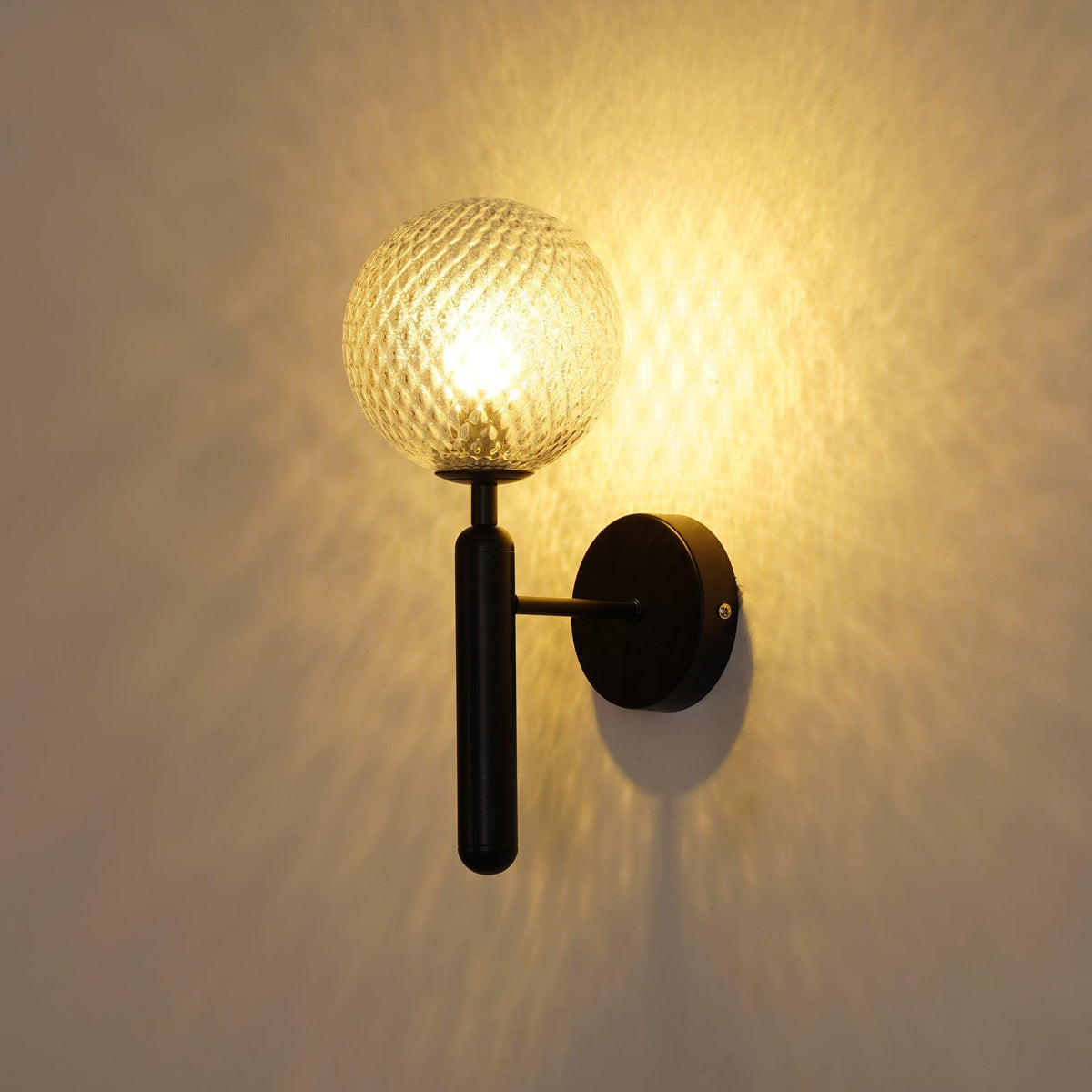 Lit warmly, this black Clérachic wall light's dimpled clear glass globe casts a strong glow across the wall behind it.