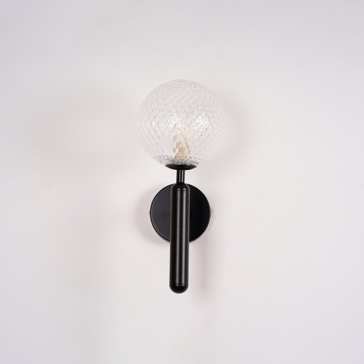 Straight-on and unlit, this black Clérachic wall light's dimpled clear glass globe stands against a plain white wall.