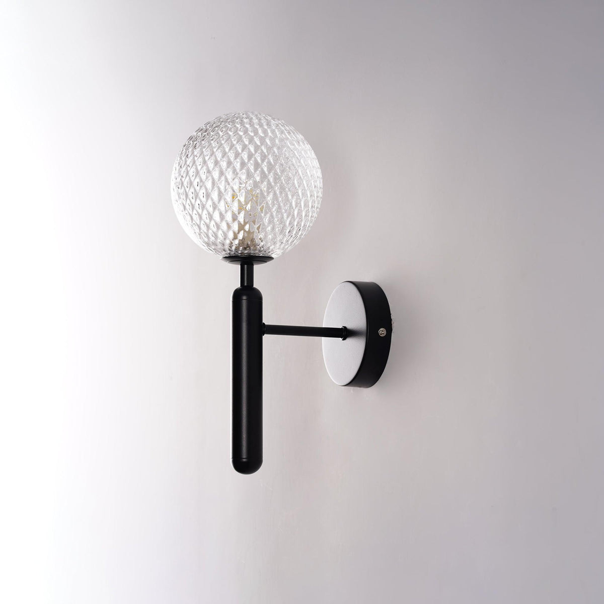 Unlit, this black Clérachic wall light shows its dimpled clear glass globe at an angle against a light grey wall.