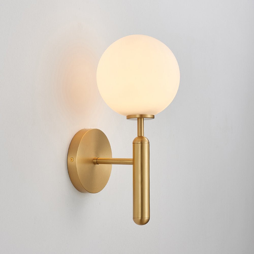 Seen at an angle, this brass Clérachic wall light shows its lit white glass globe against a plain grey wall.