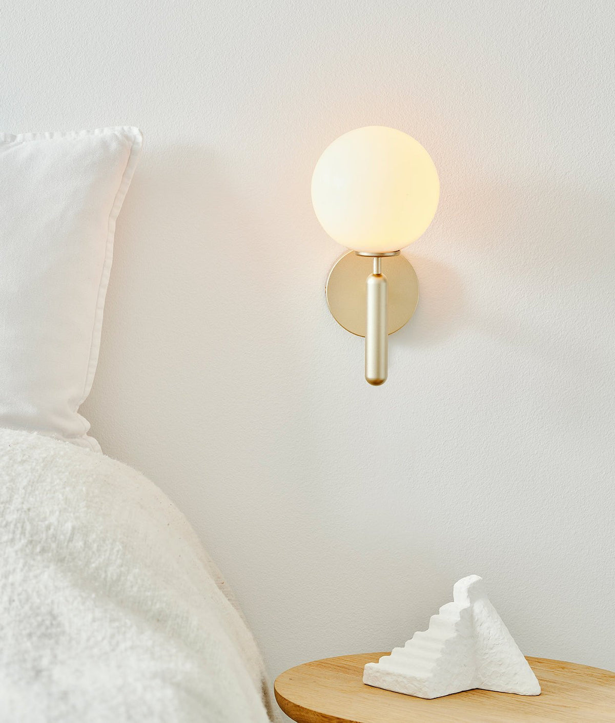 This Clérachic wall light in brushed brass glows above a bedside table with a folded pillow and a small white sculpture.