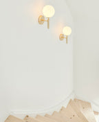 Two brass Clérachic wall lights with white glass globes light a plain white stairwell corner beside pale wood steps.