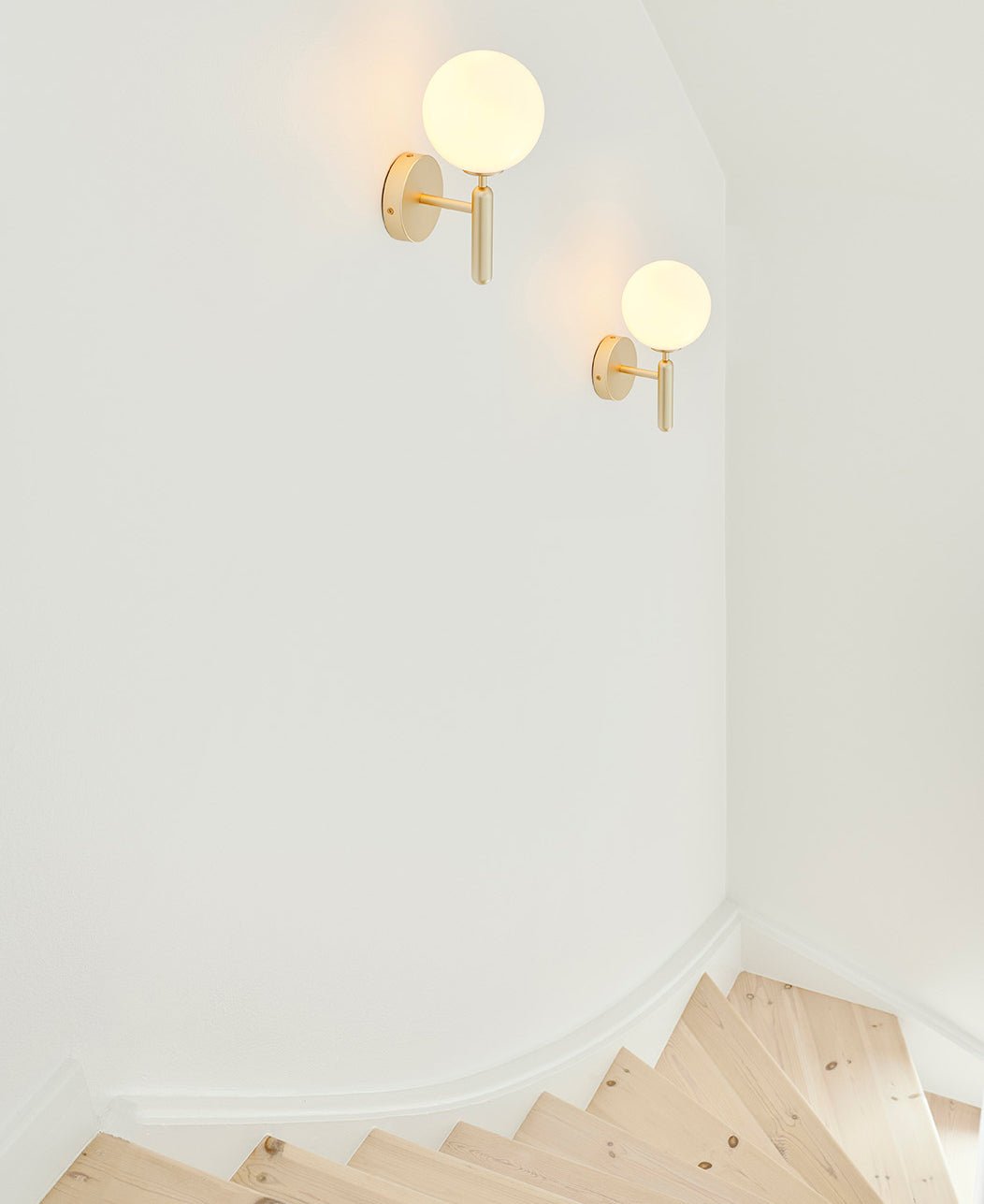 Two brass Clérachic wall lights with white glass globes light a plain white stairwell corner beside pale wood steps.