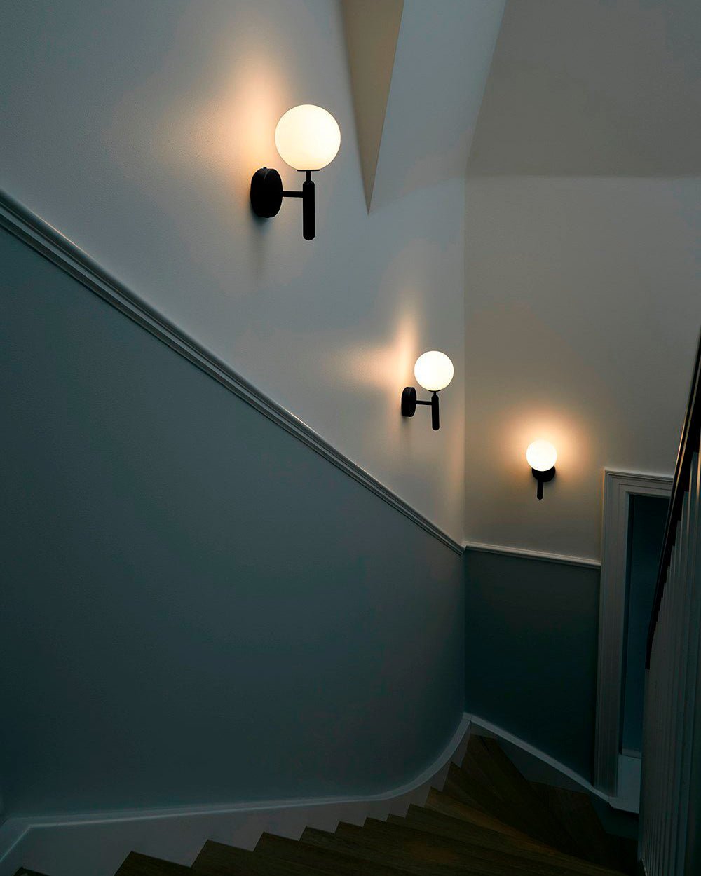 Along a stairwell painted deep teal, two black Clérachic wall lights glow near a panelled white door.