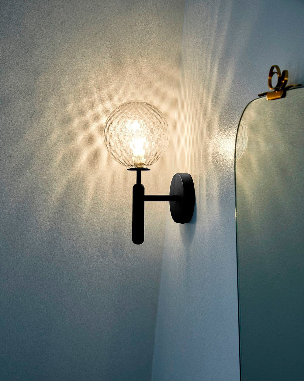 Close to an ornate mirror, this black Clérachic wall light casts light through its dimpled clear glass globe onto the wall behind.