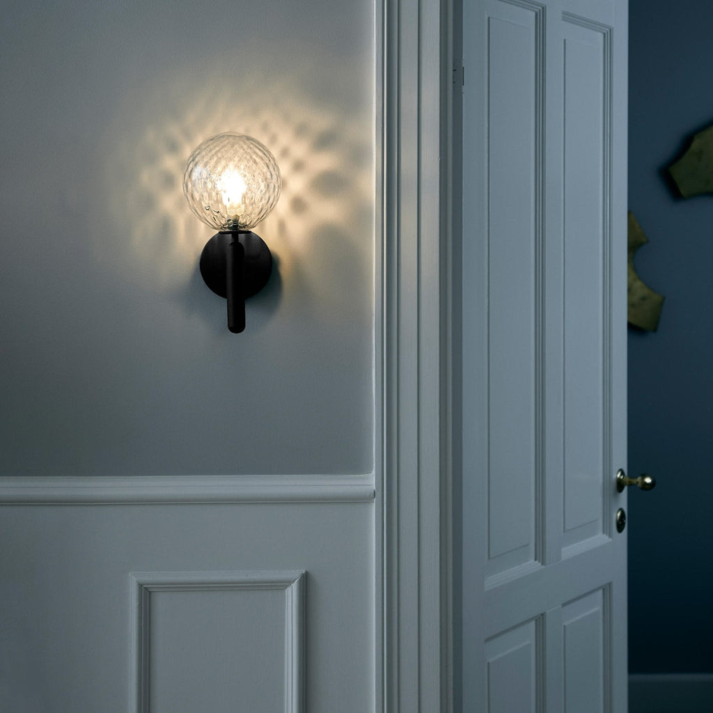 On a blue-grey panelled wall beside a white door, this black Clérachic wall light glows through its dimpled clear glass globe.
