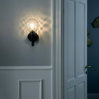 On a blue-grey panelled wall beside a white door, this black Clérachic wall light glows through its dimpled clear glass globe.