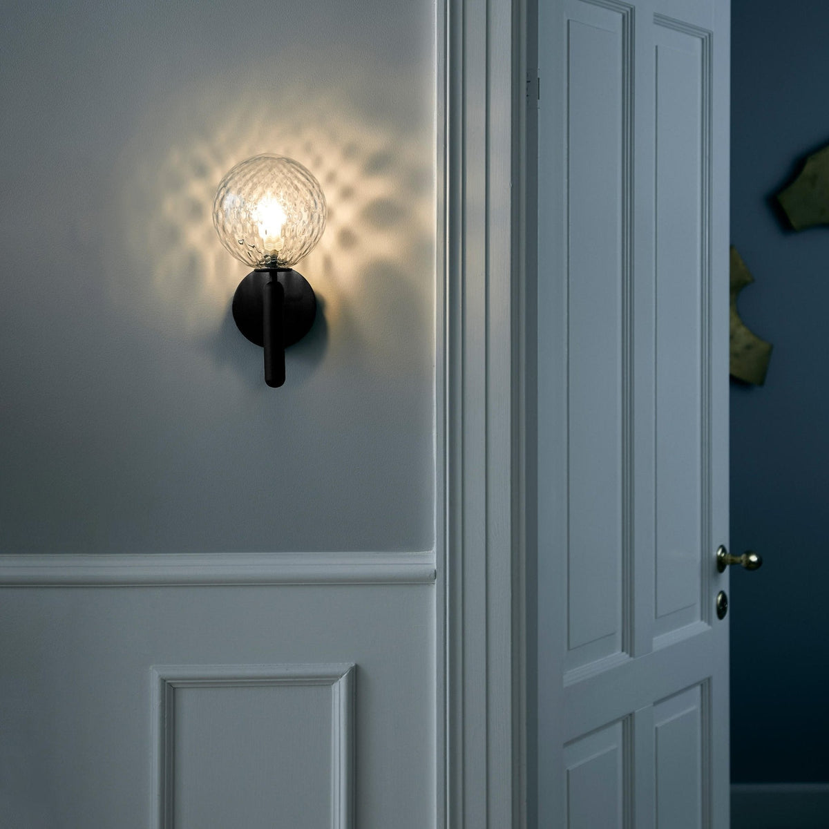 On a blue-grey panelled wall beside a white door, this black Clérachic wall light glows through its dimpled clear glass globe.